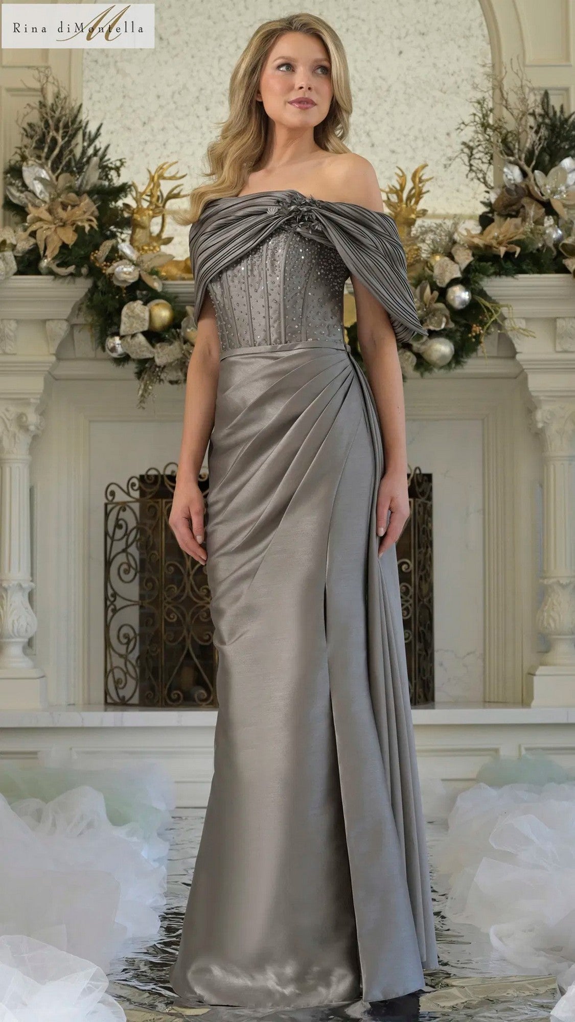 A woman in the Rina Di Montella RD3309 long formal dress in gunmetal, featuring an off-shoulder draped bodice and sheath skirt, stands before a fireplace decorated with flowers and ornaments.